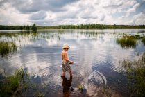 Young boy wading into peaceful lake with reflection of sky and clouds with a shovel — Stock Photo