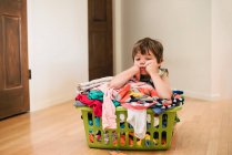 Müder Junge, der auf dem Boden sitzt und sich auf eine Wäsche lehnt, die mit Kleidung gefüllt ist — Stockfoto