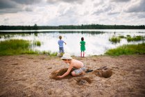 Giovane ragazzo che gioca nella sabbia vicino al lago tranquillo con riflesso del cielo e delle nuvole — Foto stock