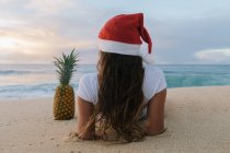 Femme portant un chapeau de Père Noël couché sur la plage à côté d'un ananas, Haleiwa, Hawaï, Amérique, États-Unis — Photo de stock