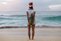 Frau mit Weihnachtsmütze, die am Strand steht und eine Ananas hinter ihrem Rücken hält, haleiwa, hawaii, america, usa — Stockfoto