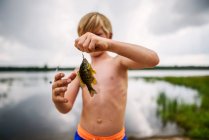 Young boy holding fish by peaceful lake with reflection of sky and clouds — Stock Photo