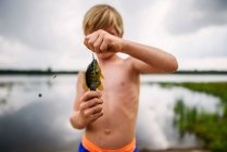 Мальчик держит рыбу у спокойного озера с отражением неба и облаков — стоковое фото