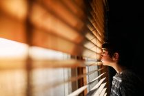 Boy standing by a window looking through blinds — Stock Photo