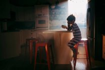 Garçon debout dans la cuisine manger son petit déjeuner dans la lumière du matin — Photo de stock