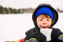 Хлопчик стоїть на снігу п'є гарячий шоколад — стокове фото
