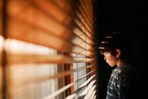 Junge steht am Fenster und schaut durch Jalousien — Stockfoto