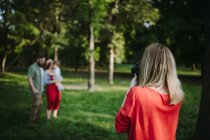 Mujer de pie en el parque tomando una foto de una familia con un niño - foto de stock