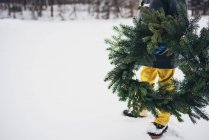Imagen recortada de Niño de pie en la nieve sosteniendo una corona de Navidad - foto de stock