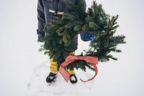 Imagen recortada de Niño de pie en la nieve sosteniendo una corona de Navidad - foto de stock