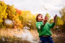 Menino de pé em um campo jogando flores de milkweed no ar — Fotografia de Stock