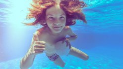 Улыбающийся мальчик под водой в бассейне держит мяч и делает большой палец вверх — стоковое фото