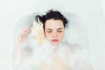 Girl lying in a bubble bath listening to a conch shell — Stock Photo