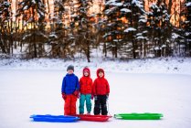 Tres niños de pie en la nieve con sus trineos - foto de stock