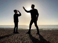 Silhouette of a Father and son standing on beach high fiving, Southsea, Hampshire, Reino Unido - foto de stock