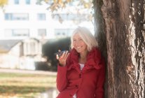 Усміхнена жінка, що схилилася до дерева, розмовляючи на мобільному телефоні (Німеччина). — стокове фото