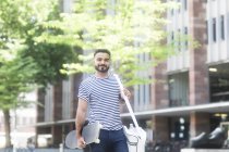 Uomo sorridente che cammina per la strada portando uno skateboard — Foto stock