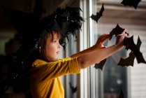 Девушка в шляпе ведьмы, приклеивающая летучую мышь украшения на окно, США — стоковое фото