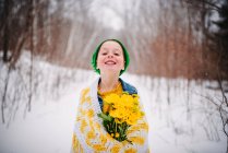 Ragazza sorridente avvolta in una coperta in piedi nella neve che tiene un mazzo di fiori gialli — Foto stock