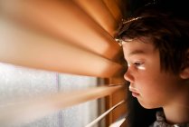 Хлопчик стоїть біля вікна, дивлячись через венеціанські штори — стокове фото