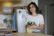Усміхнена жінка, що схиляється до горщикової рослини на кухні — стокове фото