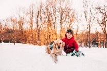 Niño sentado en la nieve con su perro golden retriever - foto de stock