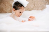Girl sitting in a bubble bath with her doll — Stock Photo