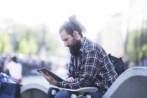 Hombre sentado en un banco apoyado en su monopatín usando una tableta digital - foto de stock