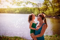 Madre de pie junto a un lago llevando a su hija - foto de stock