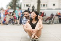 Couple souriant assis sur une planche à roulettes dans une place de la ville — Photo de stock