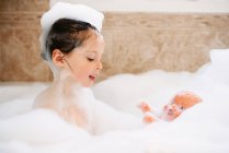 Дівчина сидить у бульбашковій ванні, граючи зі своєю лялькою — стокове фото