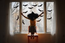 Girl wearing a witches hat kneeling on a chair by a window decorated with bats, United States — Stock Photo