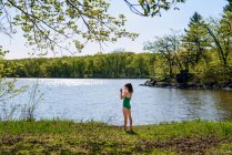Chica de pie junto a un lago en su traje de baño poniéndose sus gafas de sol - foto de stock