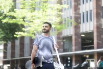 Smiling man walking down the street carrying a skateboard — Stock Photo