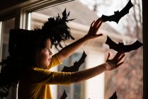 Девушка в шляпе ведьмы, приклеивающая летучую мышь украшения на окно, США — стоковое фото