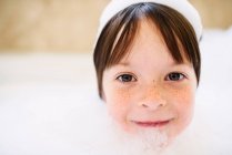 Portrait d'une fille souriante assise dans un bain moussant avec des savons sur la tête — Photo de stock