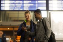 Couple standing by an arrival departure board using a mobile phone — Stock Photo