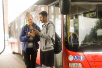 Casal em pé por um ônibus olhando para um telefone celular — Fotografia de Stock