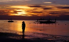 Woman walking along beach at sunset, sea water and boats — Stock Photo