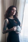 Frau steht am Fenster und hält eine Teekanne — Stockfoto