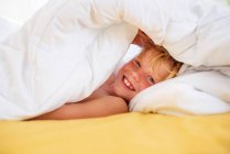 Улыбающийся мальчик в постели прячется под одеялом — стоковое фото