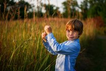 Portrait of a smiling boy standing in a field at sunset picking long grass, United States — Stock Photo