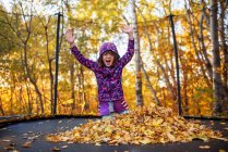 Smiling girl with her arms in the air next to a stack of autumn leaves on a trampoline, United States — Stock Photo