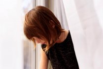 Portrait of a girl standing behind a curtain looking out of a window — Stock Photo