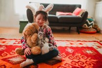 Portrait d'une fille portant des oreilles de lapin Assis sur le sol tenant des jouets mous — Photo de stock