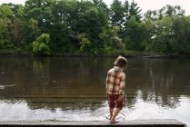 Niño parado junto a un río sumergiendo su dedo del pie en el agua, Estados Unidos - foto de stock