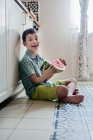 Улыбающийся мальчик, сидящий на кухонном полу с куском арбуза — стоковое фото