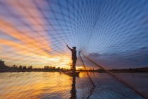 Silhouette di un pescatore in piedi su una barca a gettare una rete da pesca al tramonto, Thailandia — Foto stock