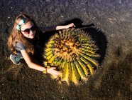 Chica sonriente agachada junto a un gran cactus redondo, Lanzarote, Islas Canarias, España - foto de stock