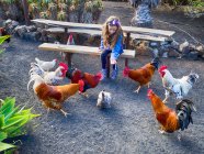 Girl sitting on a bench feeding a group of roosters and a hen, Lanzarote, Canary Islands, Spain — Stock Photo